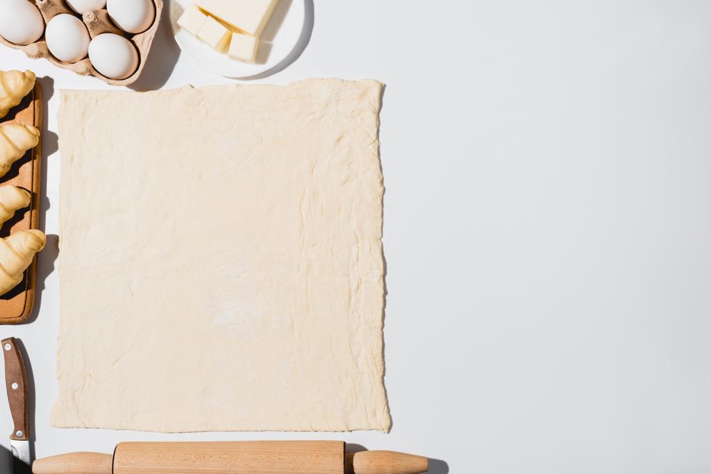 Draufsicht auf frische Croissants auf Holzschneidebrett in der Nähe von rohem Teig, Messer, Nudelholz, Butter und Eiern auf weißem Hintergrund - Foto, Bild