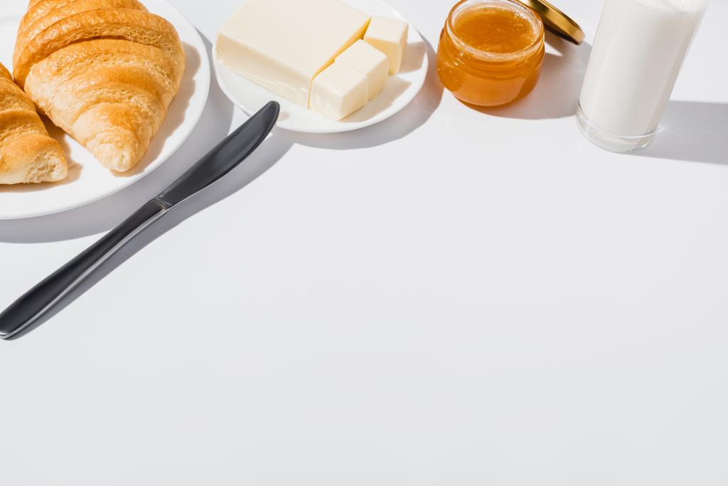 вкусные свежие круассаны на тарелке возле стакана молока, масло с ножом и варенье на белом фоне
 - Фото, изображение
