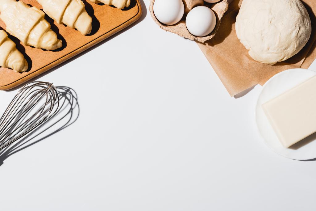 vista superior de croissants frescos na placa de corte de madeira perto da massa crua, do whisk do balão, da manteiga e dos ovos no fundo branco
 - Foto, Imagem