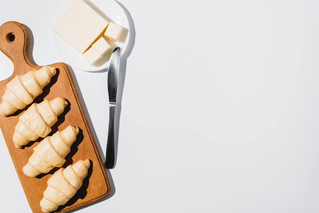vrchní pohled na čerstvé syrové croissanty na dřevěné řezací desce u másla na talíři s nožem na bílém pozadí - Fotografie, Obrázek