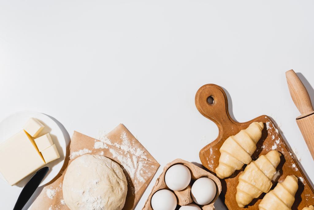 vue de dessus des croissants frais sur planche à découper en bois près de pâte crue, couteau, rouleau à pâtisserie, beurre et oeufs sur fond blanc
 - Photo, image