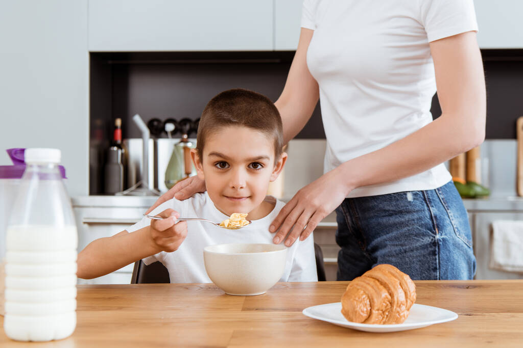Enfoque selectivo del niño mirando a la cámara mientras come cereales cerca de la madre en la cocina
  - Foto, imagen