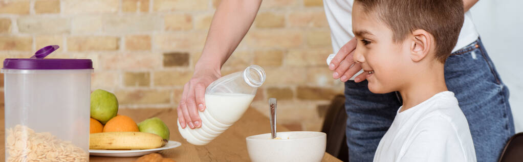 Πανοραμική κατεύθυνση της μητέρας ρίχνει γάλα στα δημητριακά κοντά χαμογελαστός γιος στο τραπέζι στην κουζίνα  - Φωτογραφία, εικόνα