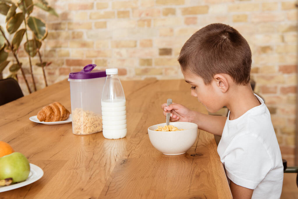 Vue latérale du garçon mangeant des céréales près du lait, croissant et fruits sur la table dans la cuisine
  - Photo, image