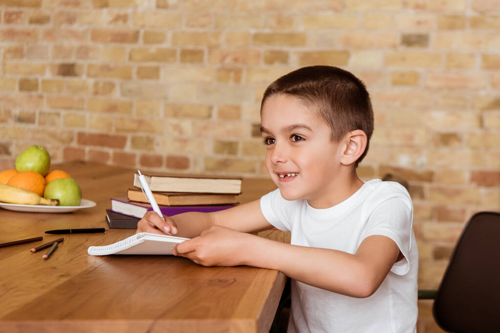 Bambino sorridente che scrive su taccuino vicino a libri e frutta sul tavolo
  - Foto, immagini