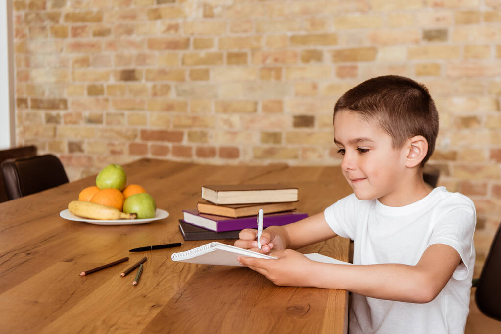 Junge schreibt auf Schreibheft neben Büchern und Bleistift auf Tisch  - Foto, Bild