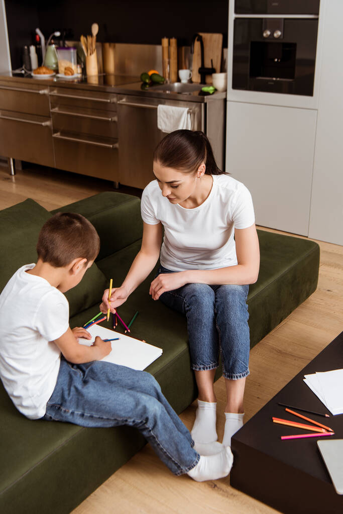 ソファの上に描かれた息子の近くに色鉛筆を保持する母親の選択的な焦点  - 写真・画像