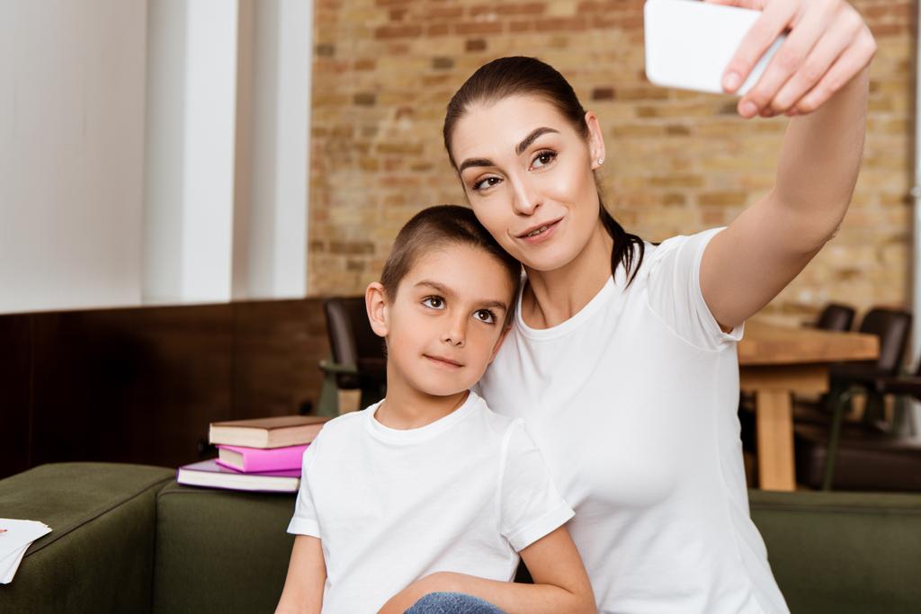 Selektivní zaměření matky a roztomilý syn přičemž selfie s smartphonem v blízkosti knih na gauči  - Fotografie, Obrázek