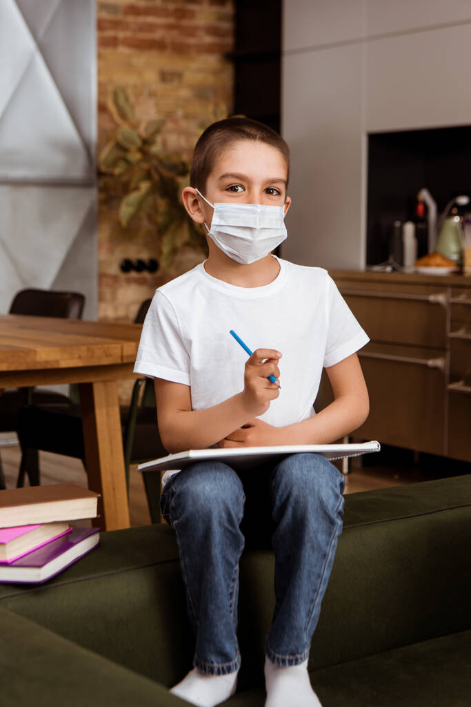 Selectieve focus van kind in medisch masker met schetsboek en potlood in de buurt van boeken op de bank  - Foto, afbeelding
