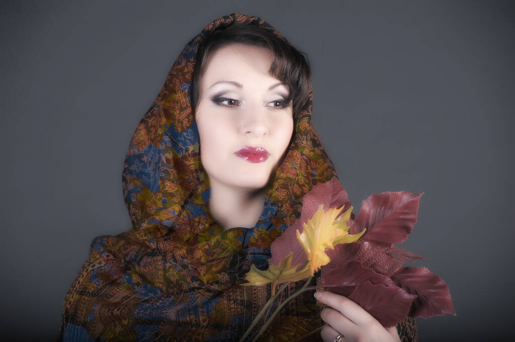 beautiful Russian brunette woman with a scarf on her head and autumn leaves in her hands, photo in the studio on a gray background - Photo, Image