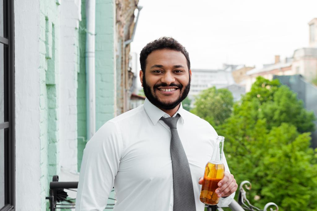 lächelnder und bärtiger Afroamerikaner mit einer Flasche Bier auf dem Balkon  - Foto, Bild