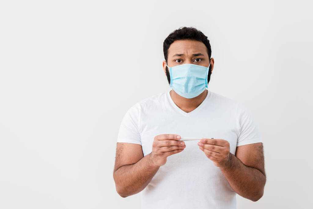 nemocný africký Američan v lékařské masce drží digitální teploměr v blízkosti bílé stěny  - Fotografie, Obrázek