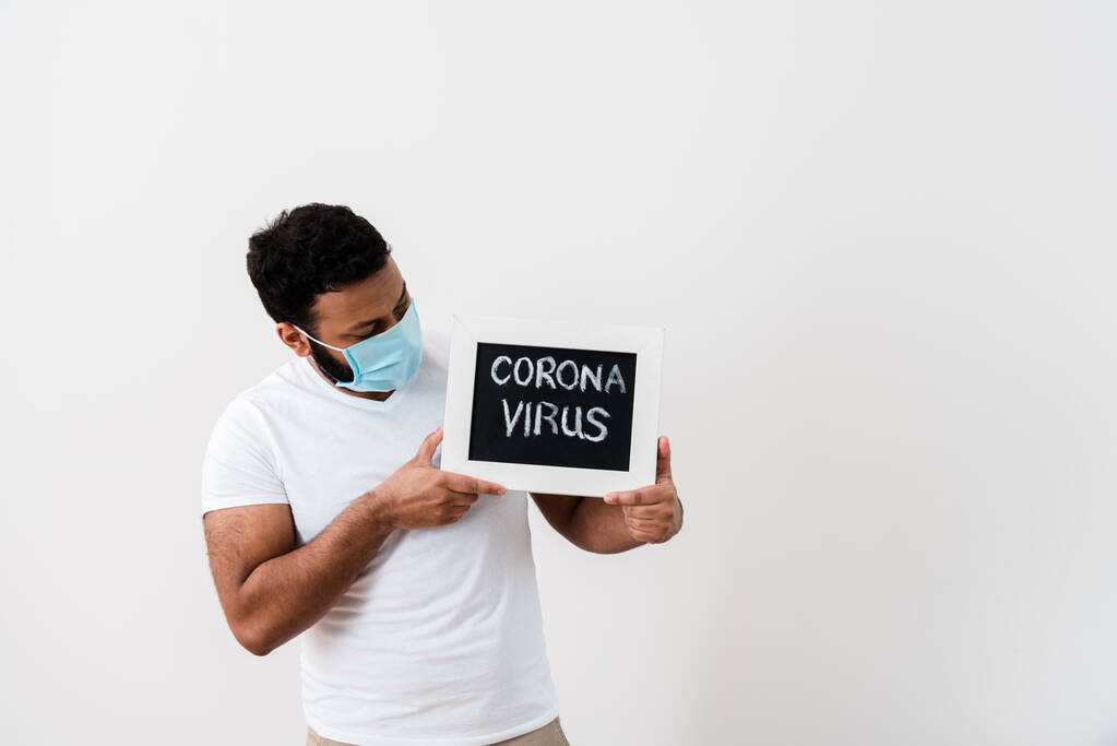 青い医療マスクのアフリカ系アメリカ人男性白い壁の近くにコロナウイルスの文字で黒板を見て  - 写真・画像