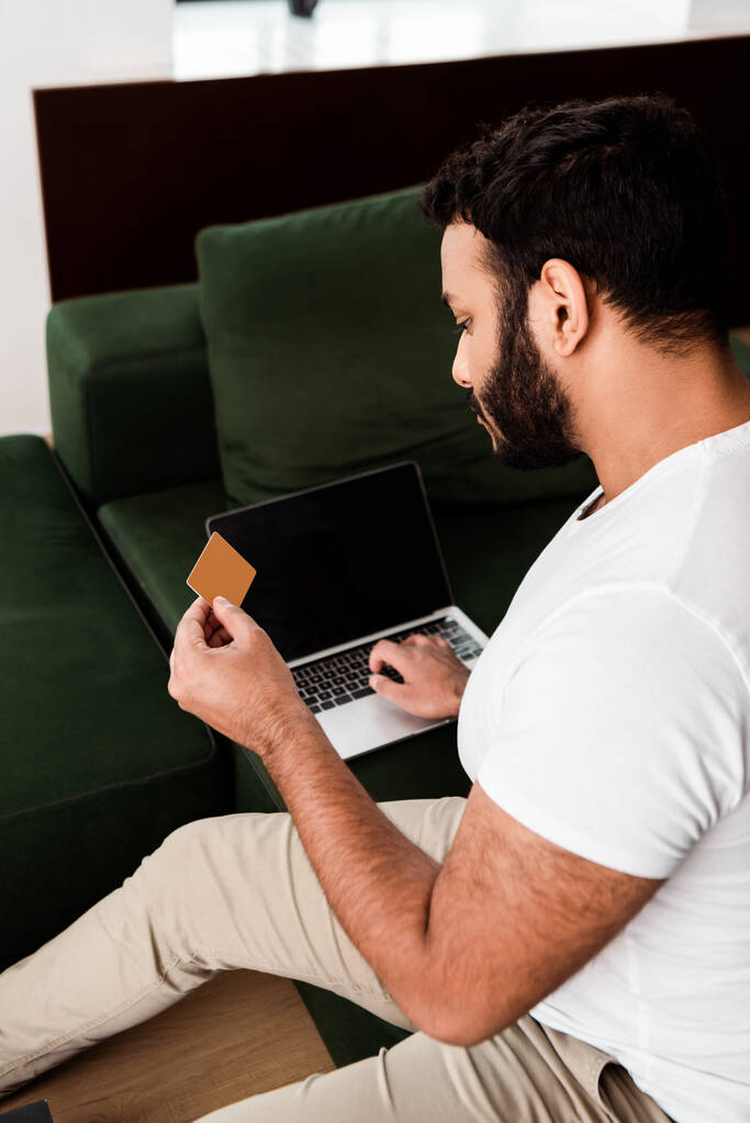 selective focus of bearded african american man holding credit card near laptop with blank screen while online shopping in living room  - Photo, Image