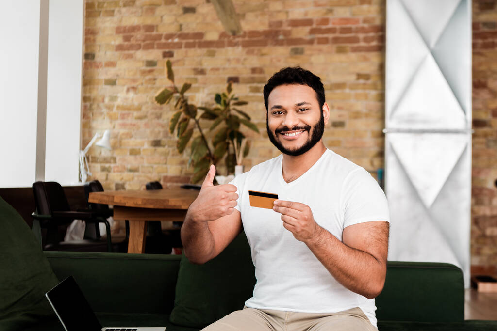 ευτυχισμένος αφρικανός Αμερικανός άνδρας που κρατά την πιστωτική κάρτα κοντά στο lap-top με την κενή οθόνη ενώ δείχνει τον αντίχειρα επάνω  - Φωτογραφία, εικόνα
