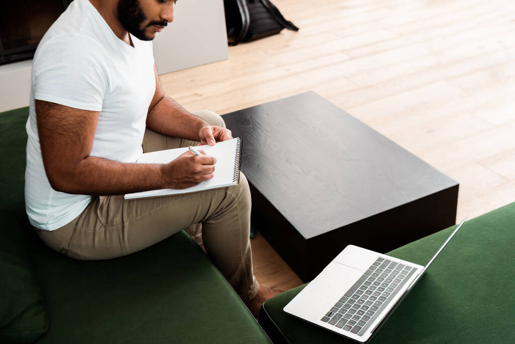 bijgesneden weergave van bebaarde Afrikaans-Amerikaanse man schrijven in notebook in de buurt van laptop, studie online concept - Foto, afbeelding