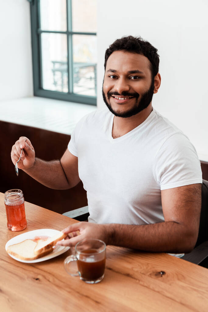 onnellinen afrikkalainen amerikkalainen mies pitää lusikka lähellä purkki makea hilloa ja paahtoleipää lautasella
 - Valokuva, kuva