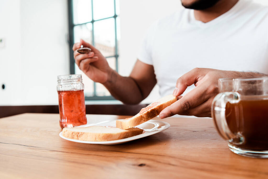 bijgesneden uitzicht van Afrikaanse Amerikaanse man die lepel in de buurt van pot met zoete jam en toast brood op bord - Foto, afbeelding