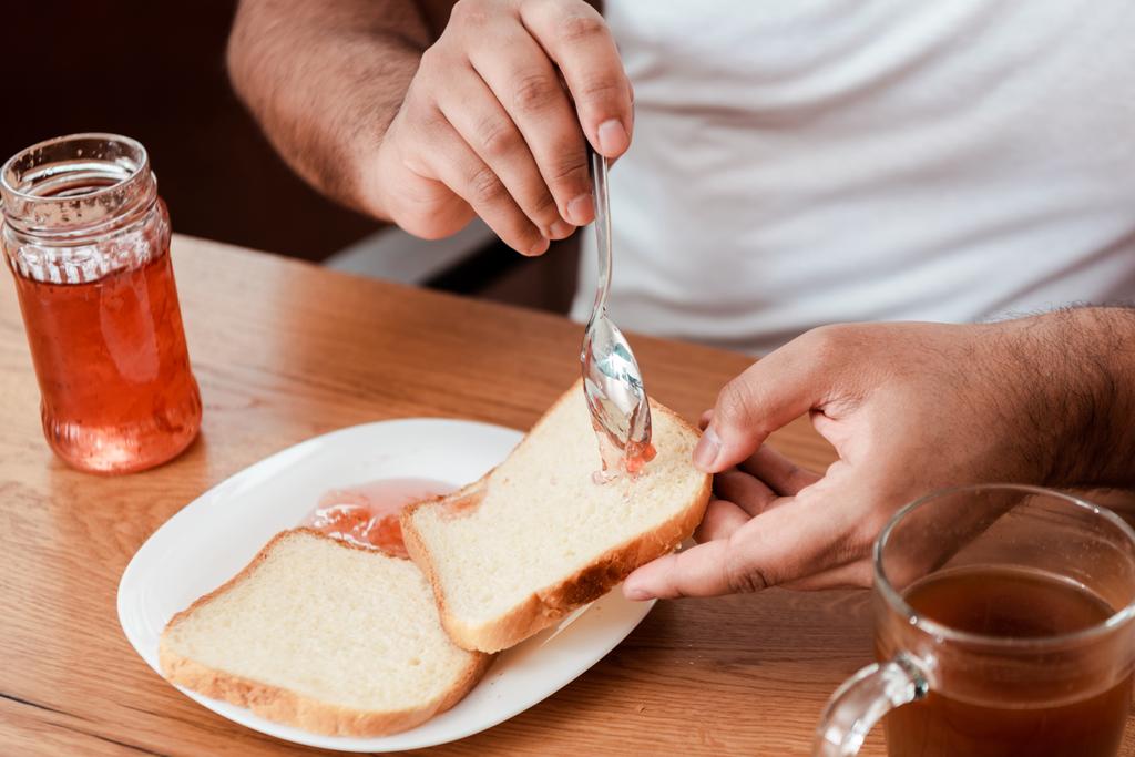 vista recortada del hombre afroamericano sosteniendo cuchara con mermelada dulce cerca de pan tostado en el plato
 - Foto, imagen
