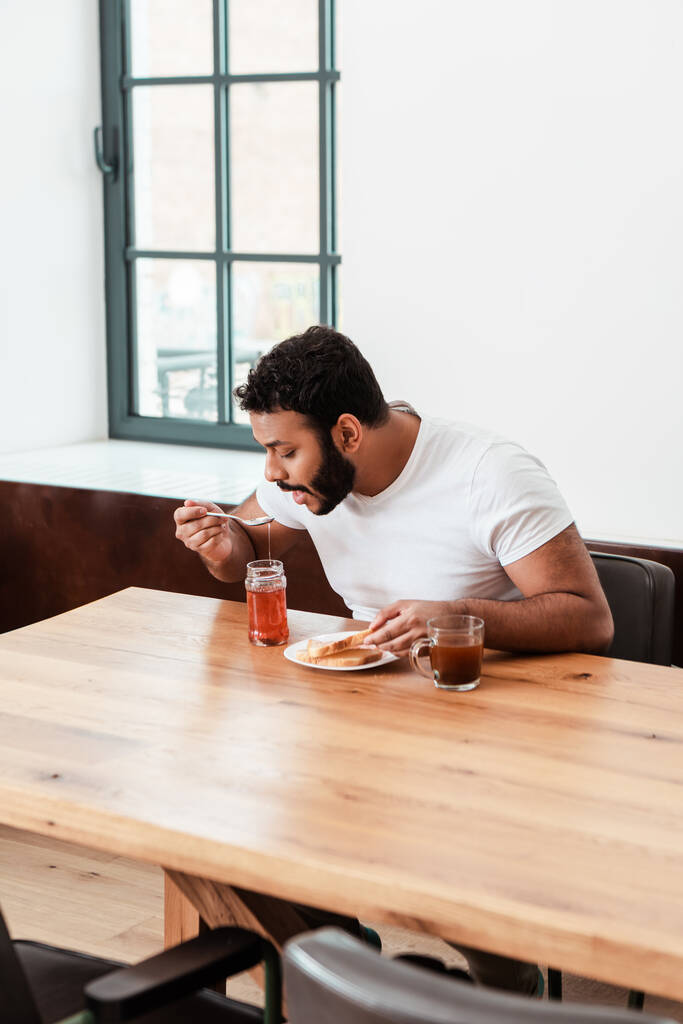 uomo africano americano che tiene cucchiaio con marmellata dolce vicino alla bocca aperta e pane tostato sul piatto e tazza di caffè
 - Foto, immagini