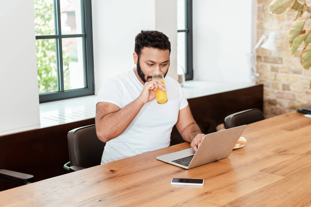 アフリカ系アメリカ人のフリーランスは、スマートフォンの近くのノートパソコンを空の画面で使いながら新鮮なオレンジジュースを飲む - 写真・画像