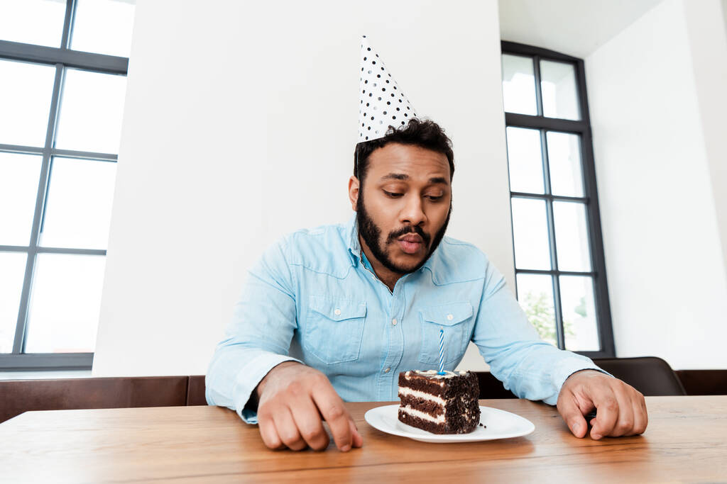 african american man in party cap celebrating birthday alone and blowing out candle on birthday cake - Photo, Image