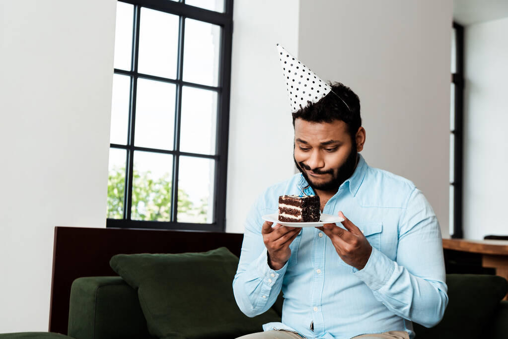 displeased african american man in party cap holding plate with birthday cake - Photo, Image