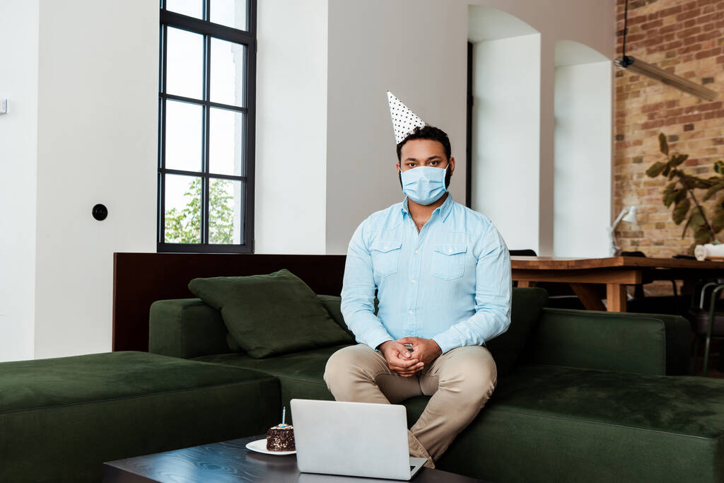 african american man in party cap and medical mask near laptop and birthday cake - Photo, Image
