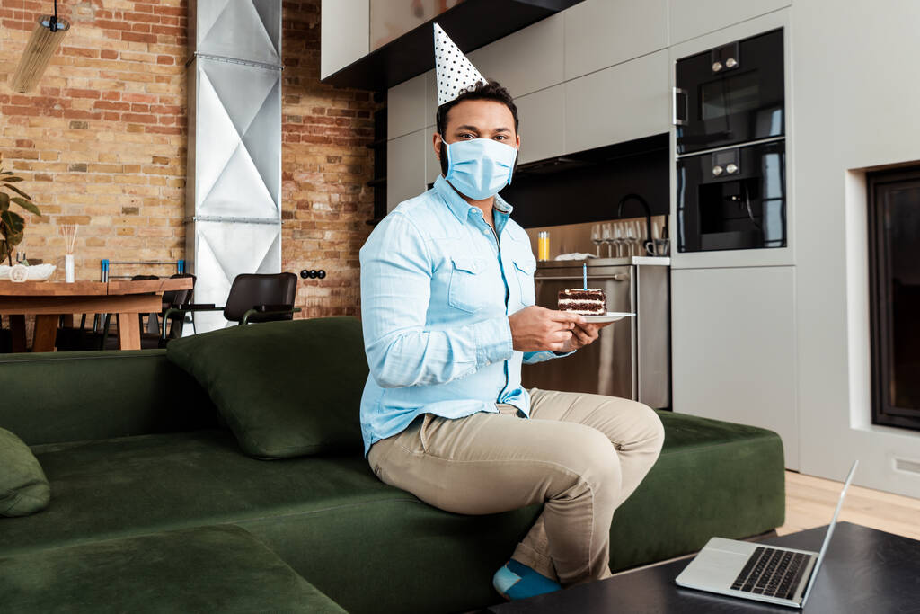 Afrikaan amerikaanse man met medisch masker en feestmuts holding plaat met smakelijke verjaardagstaart in de buurt van laptop in de woonkamer - Foto, afbeelding