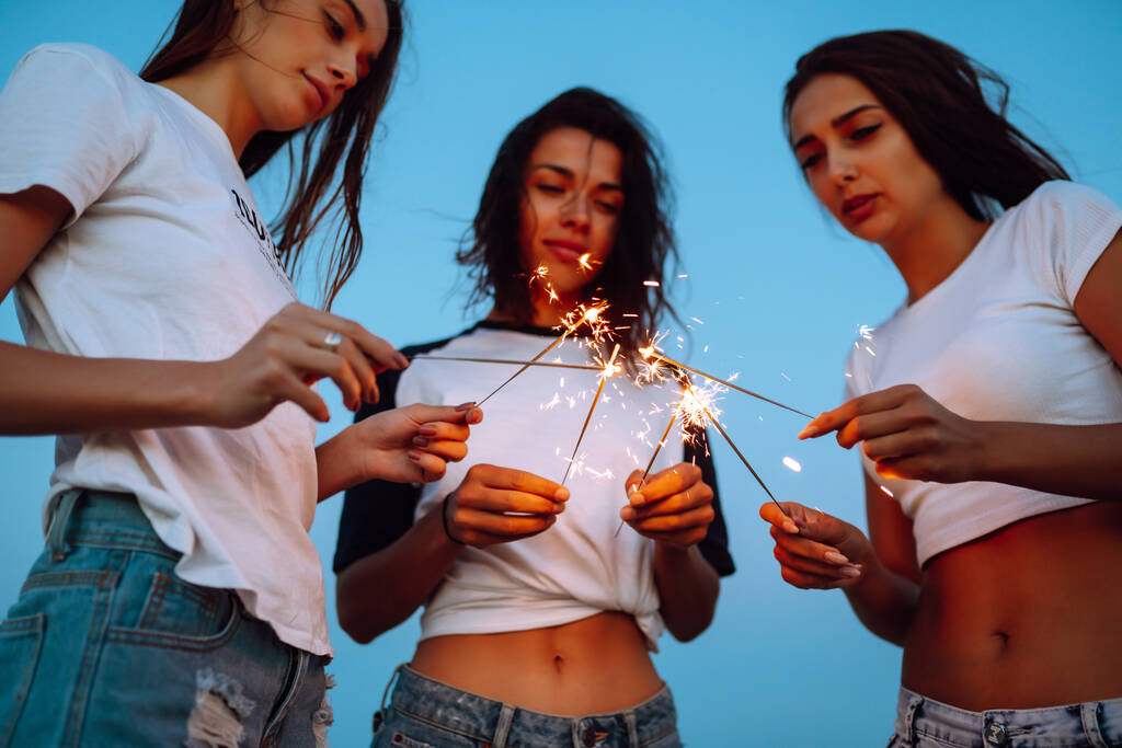 Błyskotki w rękach młodych dziewczyn na plaży. Trzy dziewczyny cieszące się imprezą na plaży z ogniem. Wakacje, wakacje, relaks i koncepcja stylu życia. - Zdjęcie, obraz