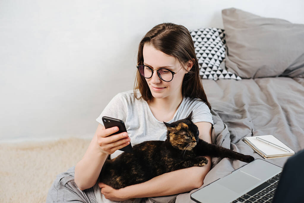 Uśmiechnięta młoda kobieta w okularach profesjonalny użytkownik siedzi na podłodze w domu trzymając kota i smartfona w ręku SMS-a. Cute girl za pomocą technologii aplikacji komórkowych nowoczesny gadżet koncepcja. - Zdjęcie, obraz