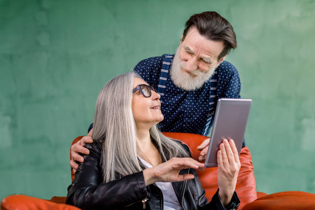 Студийный снимок счастливой пожилой семейной пары, стильных мужчины и женщины, смотрящих друг на друга во время просмотра Интернета или использования приложений на планшете i-pad и разговаривающих друг с другом. Женщина сидит в красном кресле
 - Фото, изображение