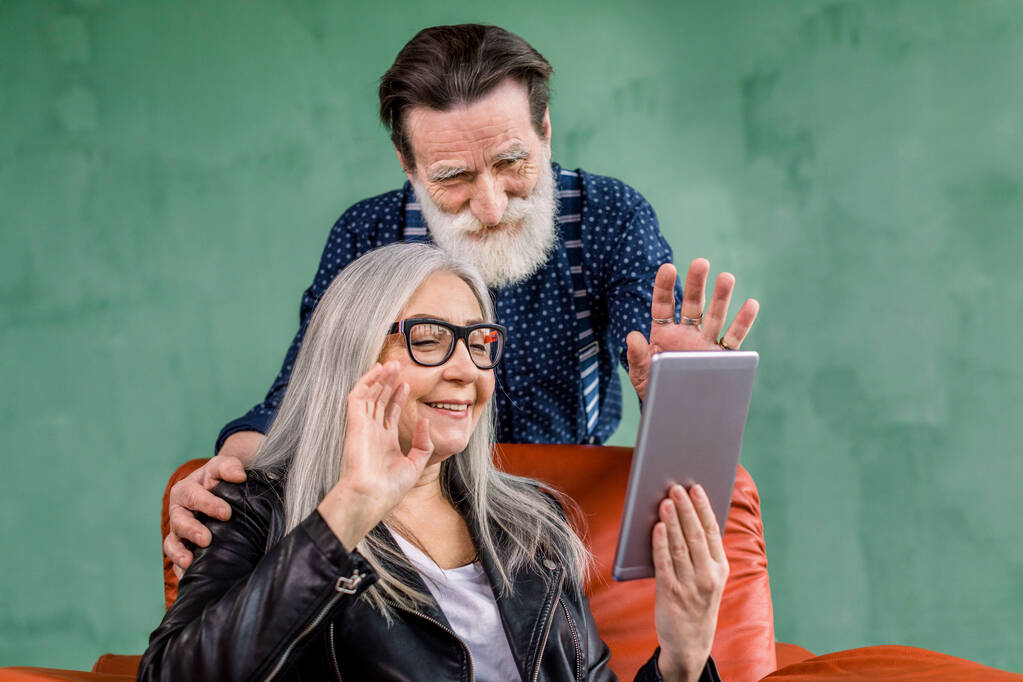 Αρκετά χαρούμενη 60-70 γκρίζα μαλλιά κυρία κάθεται σε μαλακή κόκκινη καρέκλα και κρατώντας tablet για βιντεοκλήση, ενώ όμορφος γενειοφόρος σύζυγος ακουμπά στην καρέκλα και την αγκαλιάζει, χαιρετώντας μαζί - Φωτογραφία, εικόνα
