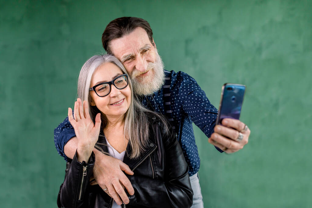 Portret atrakcyjnej pary seniorów, stojącej w domu przed zieloną ścianą i rozmawiającej z przyjaciółmi przez telefon. brodaty mężczyzna obejmujący jego żonę podczas gdy ona macha ręką do aparatu telefonicznego - Zdjęcie, obraz