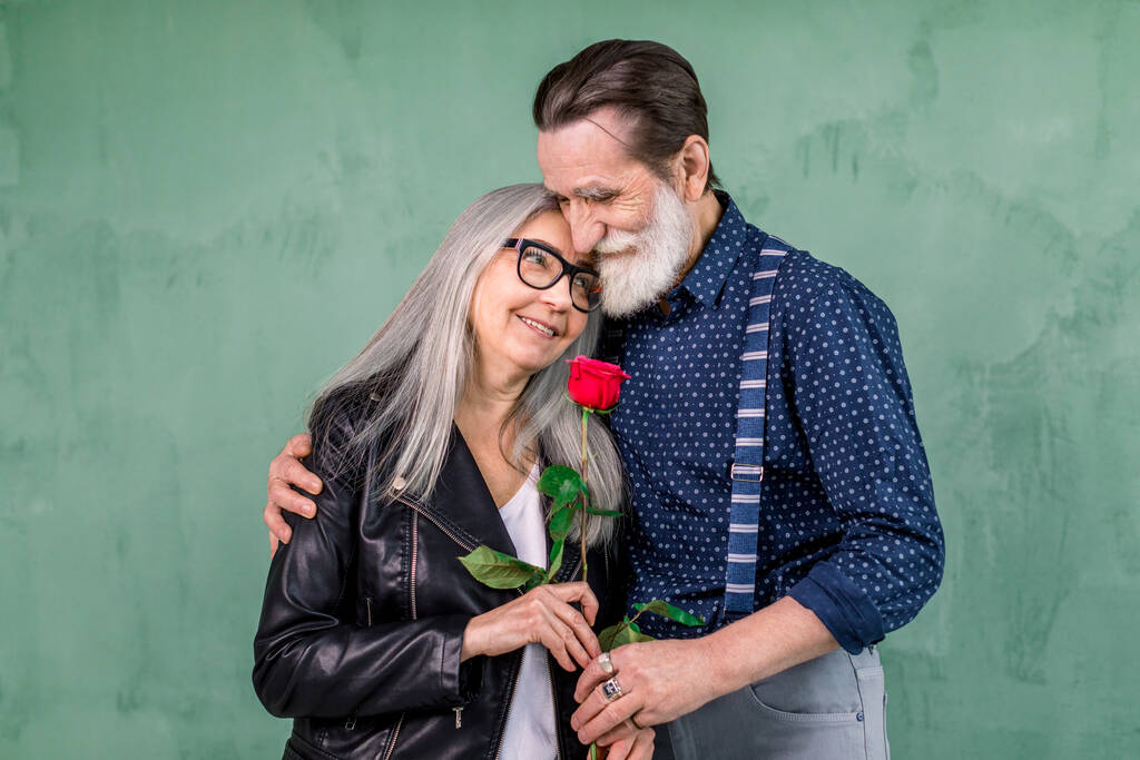 Love has no age. Romantic senior couple celebrating Saint Valentines Day. Beautiful woman and handsome man enjoying spending time together, embracing and holding red rose - Photo, Image