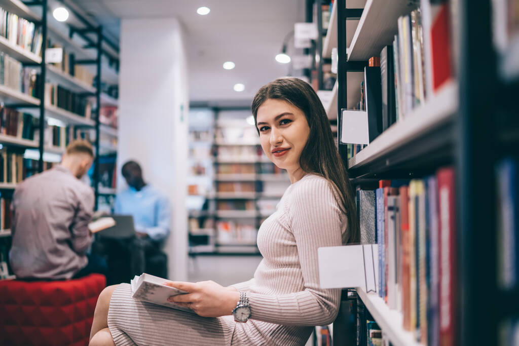 Bela senhora de vestido sentado com pernas cruzadas e desfrutando de história no livro enquanto arrefece na biblioteca e olhando para a câmera  - Foto, Imagem