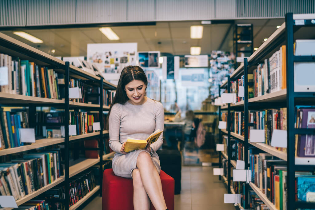Trendige junge Frau im Kleid lächelt und liest interessante Bücher, während sie auf rotem Hocker neben Bücherregalen in der modernen Bibliothek sitzt - Foto, Bild