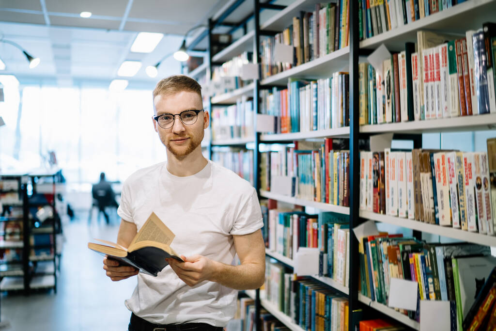 Χαμογελώντας ενηλίκων γενειοφόρος φοιτητής σε casual ρούχα και γυαλιά κρατώντας άνοιξε το βιβλίο και κοιτάζοντας την κάμερα, ενώ στέκεται στο φως σύγχρονη βιβλιοθήκη - Φωτογραφία, εικόνα