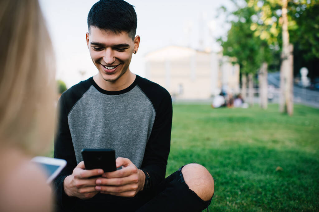 Fröhlich lächelnde zufriedene Männer, die per Smartphone SMS schreiben und soziale Medien nutzen, während sie mit Freunden auf grünem Gras im Stadtpark entspannen - Foto, Bild