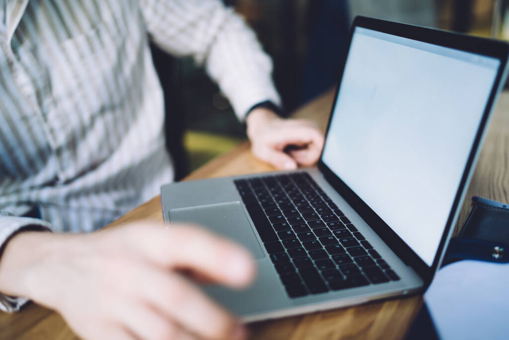 Onherkenbare kantoormedewerker in gestreept shirt zittend voor open laptop met leeg scherm op tafel op wazige achtergrond - Foto, afbeelding