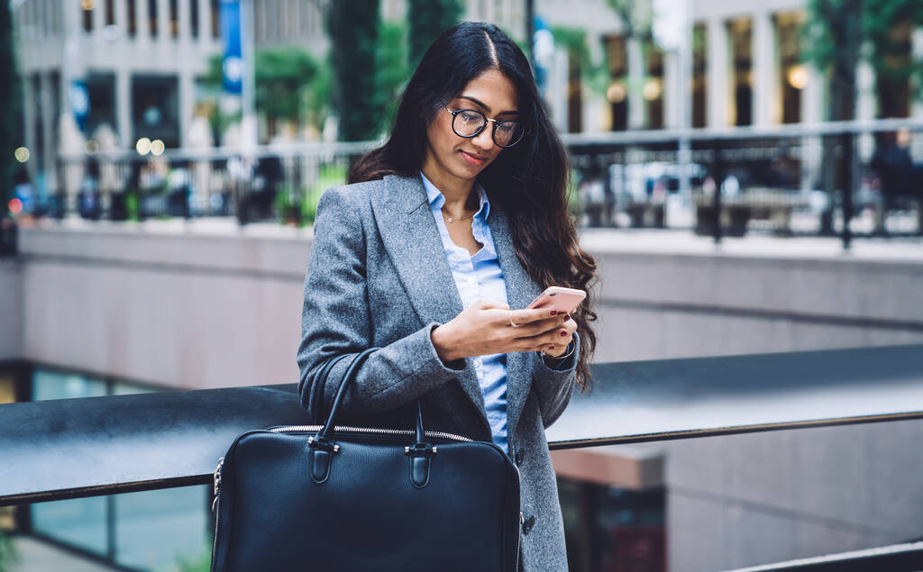 Fajna etniczna kobieta w okularach i szarym płaszczu niosąca skórzaną torbę i wiadomość na telefonie komórkowym stojącym na tle miasta - Zdjęcie, obraz