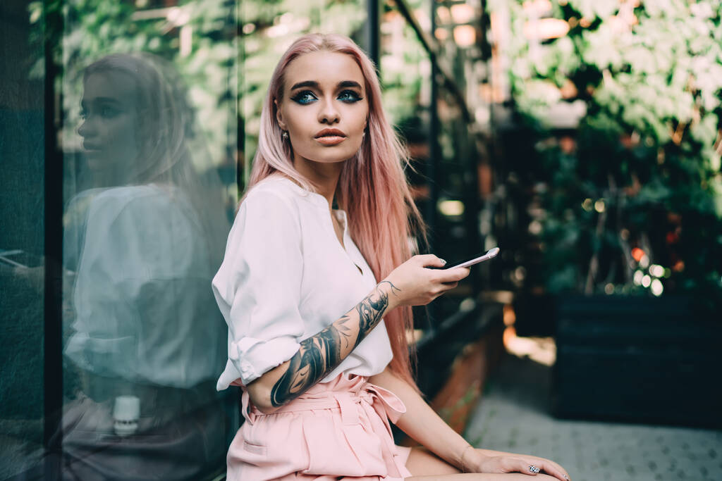 Vista lateral de mujer elegante con pelos largos de color rosa y ojos verdes mirando hacia otro lado surfeando teléfono móvil apoyado en la pared de cristal del edificio - Foto, imagen