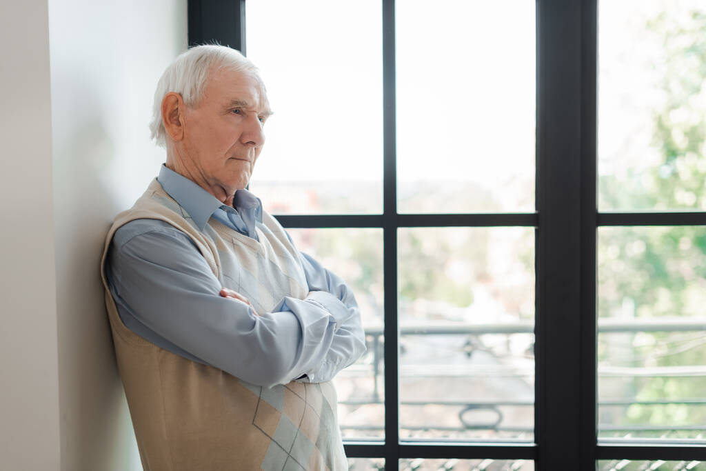 старший самотній чоловік з схрещеними руками, що стоять біля вікна під час самоізоляції
 - Фото, зображення