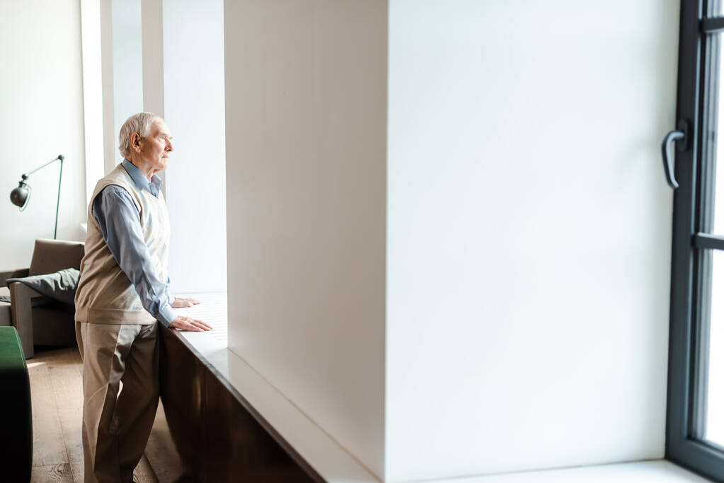 szomorú idős férfi, aki az ablak mellett áll az elszigeteltség idején - Fotó, kép