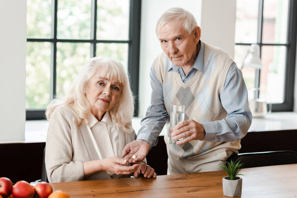ανήσυχος ηλικιωμένος σύζυγος και άρρωστη γυναίκα με χάπια και ένα ποτήρι νερό στο σπίτι κατά τη διάρκεια της καραντίνας - Φωτογραφία, εικόνα