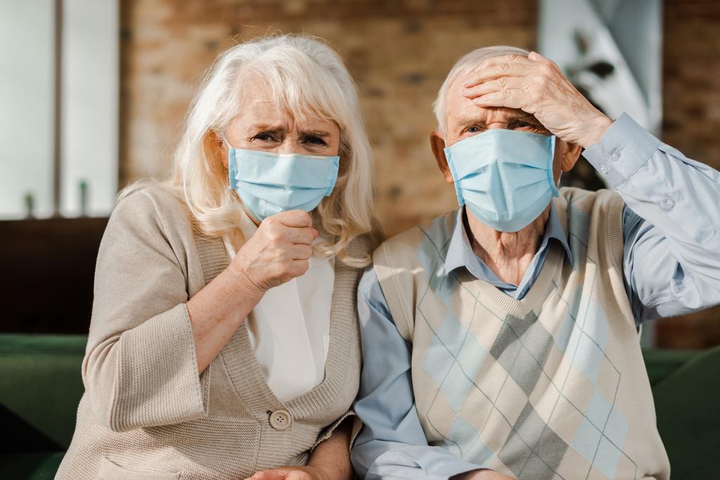 ηλικιωμένο ανήσυχο ζευγάρι με ιατρικές μάσκες κάθεται στο σπίτι κατά τη διάρκεια της απομόνωσης - Φωτογραφία, εικόνα