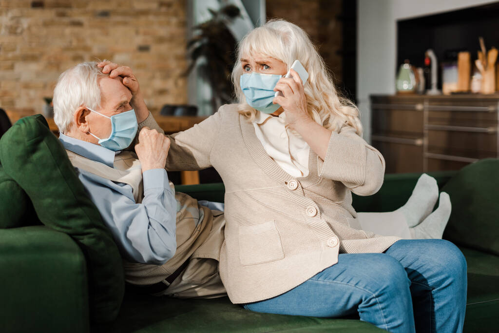 senior woman in medical mask calling doctor with smartphone while ill husband with fever lying on sofa during coronavirus epidemic  - Photo, Image
