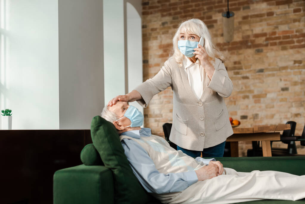 iäkäs nainen lääketieteellisessä naamiossa soittamalla lääkärille älypuhelimella, kun sairas aviomies makaa sohvalla sepelvaltimotautiepidemian aikana  - Valokuva, kuva