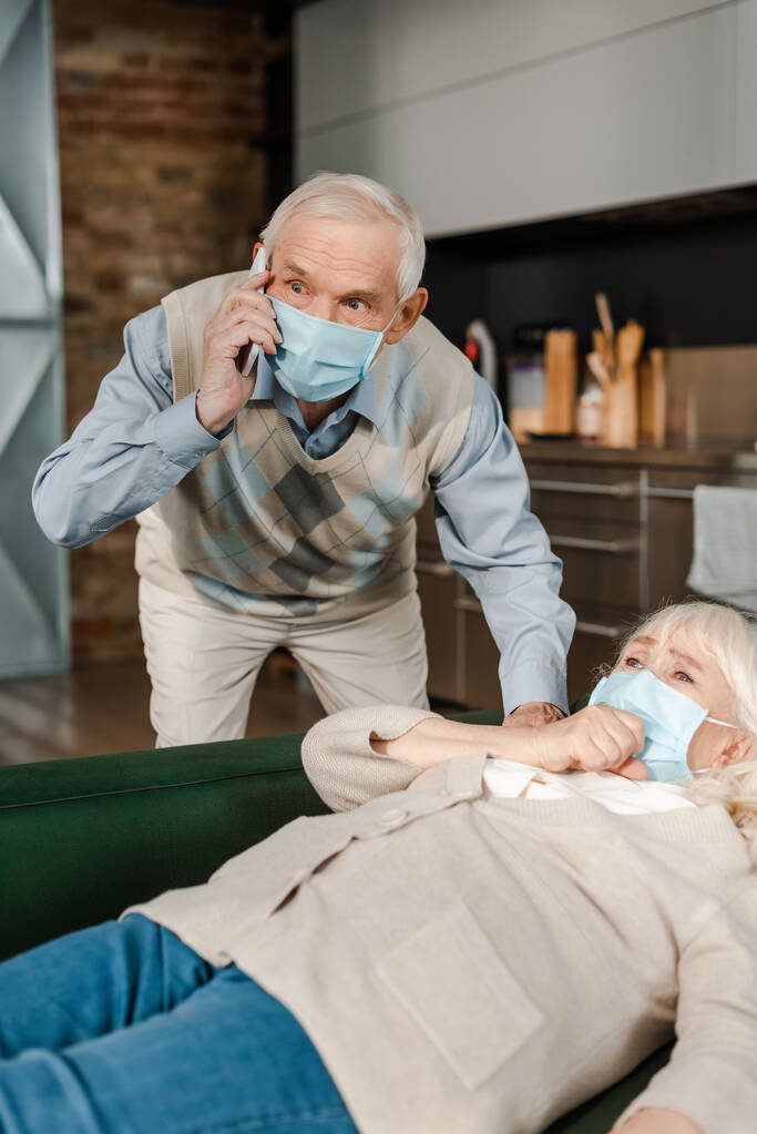 Besorgter älterer Mann in medizinischer Maske ruft Arzt mit Smartphone an, während kranke Frau während Selbstisolierung auf Sofa liegt - Foto, Bild