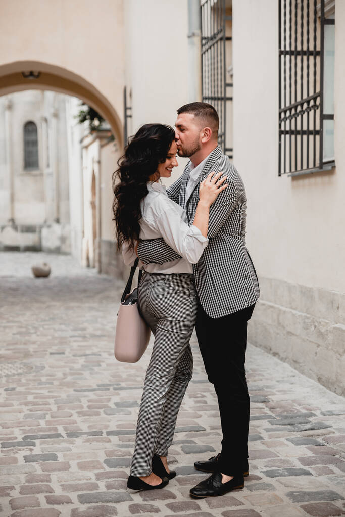 Charming girl with long brown hair and a man with a beard kissing on the background of the morning city. Newlyweds on their wedding day - Photo, Image